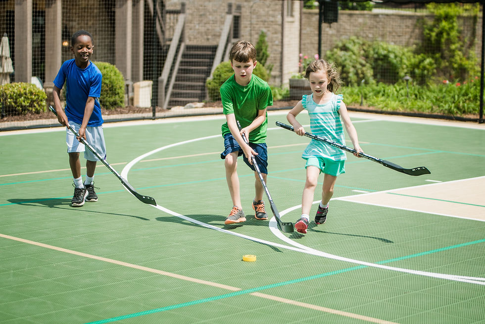 Three children play street hockey with sticks on a green outdoor court, chasing a yellow puck. They're joyful, under a sunny sky.