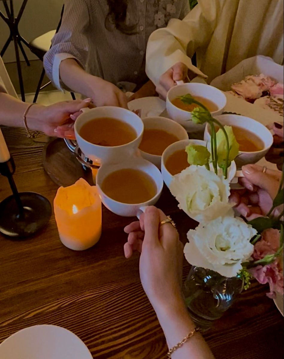 Hands raise white teacups around a wooden table with candles and flowers, creating a warm, cozy gathering mood
