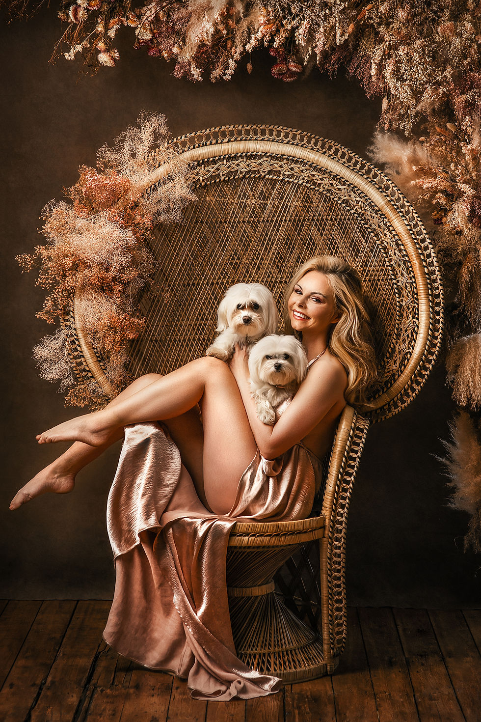 A joyful portrait of a woman sitting gracefully in a large rattan chair, surrounded by dried flowers. She is wearing a silky dress and smiling as she holds two fluffy dogs in her arms, exuding warmth and happiness against a soft, textured background