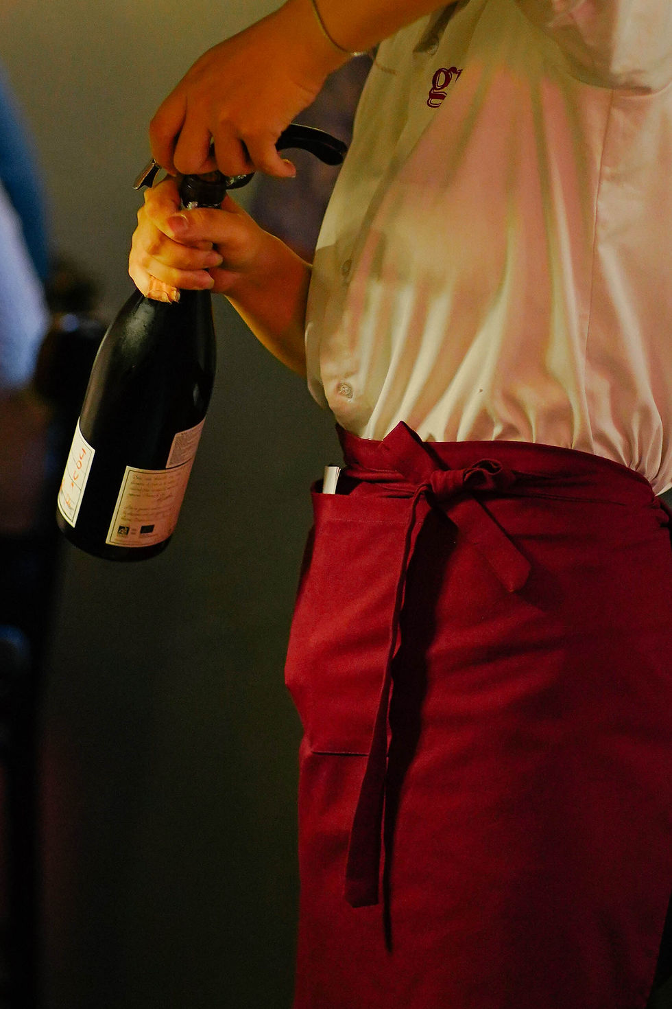 Person in red apron opening a wine bottle, wearing a white shirt with a faint logo. Neutral tone background.