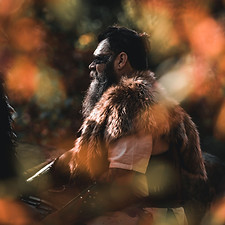 A couple kneels in a shallow, forested stream, embracing each other closely. The woman, dressed in a dark, off-the-shoulder g
