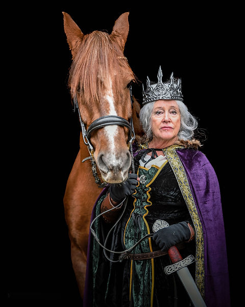 An older woman with silver hair, wearing a regal crown and a royal-style cape, sits confidently atop a chestnut horse in an