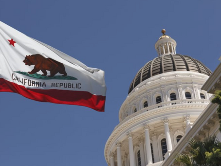 An image of the California State Capitol dome in Sacramento under a clear blue sky, with the California State Flag (the Bear Flag) waving prominently in the foreground. The image represents California Senate Bill 729 (SB 729), a landmark law signed in 2024 that mandates expanded insurance coverage for infertility treatments and IVF starting in 2026.