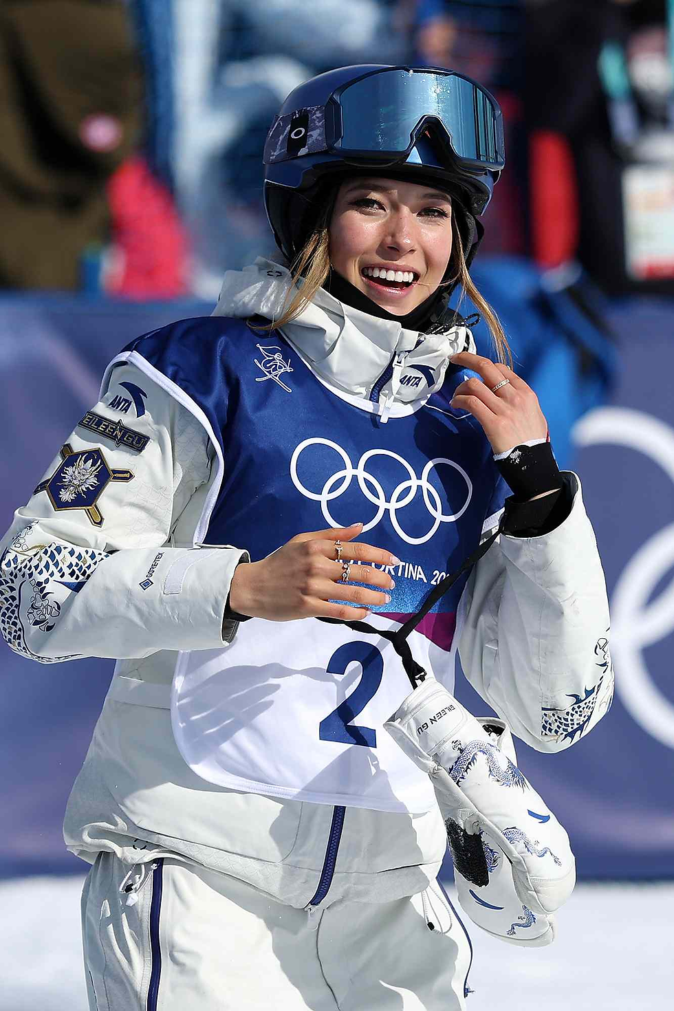 一名身穿中国队比赛服的女子自由式滑雪运动员戴着头盔和雪镜,在冬奥会赛场上微笑,胸前佩戴印有奥运五环标志的号码布。