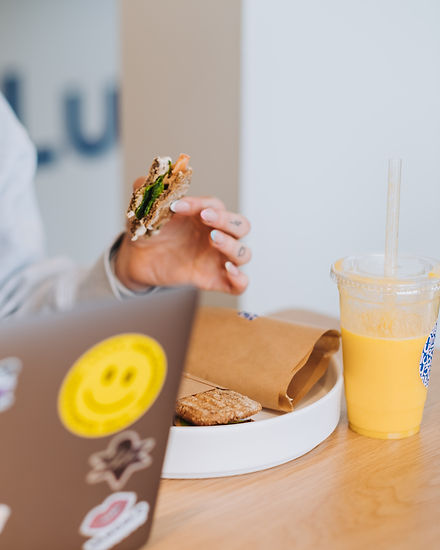 Hånd som holder en sandwich som er spist på, appelsinjuice på siden og en mac som jobbes på