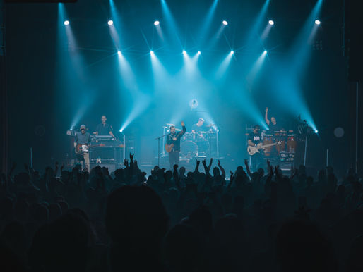 A concert photo of Umphrey's McGee performing live at Mayo Performing Arts Center in Morristown, NJ for their Sky's The Limit Tour. The photo is wide angle with the full band on stage illuminated by blue lights and a cheering crowd in front. The photo is taken by Ryan Downey Jr. for S!CK Entertainment
