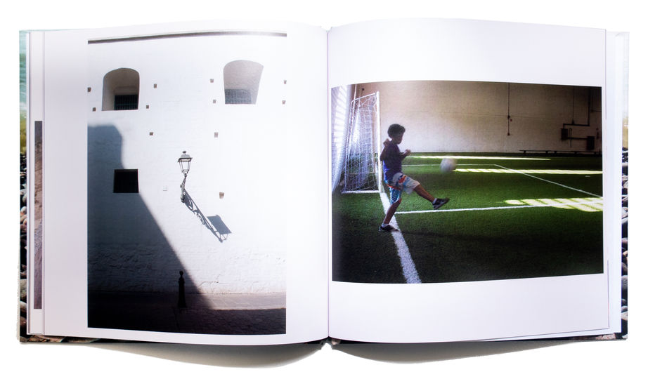 Páginas internas do livro Descanso: nas reticências do caminho, de autoria de  Maína Fantini
A imagem mostra o livro aberto, com uma fotografia em cada página.
Na página da esquerda, uma parede branca, com arcos e pequenas janelas que se distribuem pela superfície. Uma lanterna de poste projeta sombra longa e nítida, criando uma diagonal escura que domina a composição.
Na página da direita, uma criança em movimento sobre um campo de grama sintética verde, prestes a chutar uma bola em direção ao gol. A figura está em contraluz, em silhueta parcial. A luz entra lateralmente pelo fundo, criando faixas amareladas sobre o gramado.
As duas fotografias dialogam entre si pela forma, pela luz e pela sombra: a diagonal escura da lanterna, da fotografia à esquerda, ecoa no movimento do corpo da criança, da fotografia à direita, e o contraste entre claro e escuro se repete nas duas cenas, formando uma composição singular, criativa e harmoniosa dentro da proposta contemplativa do livro.
