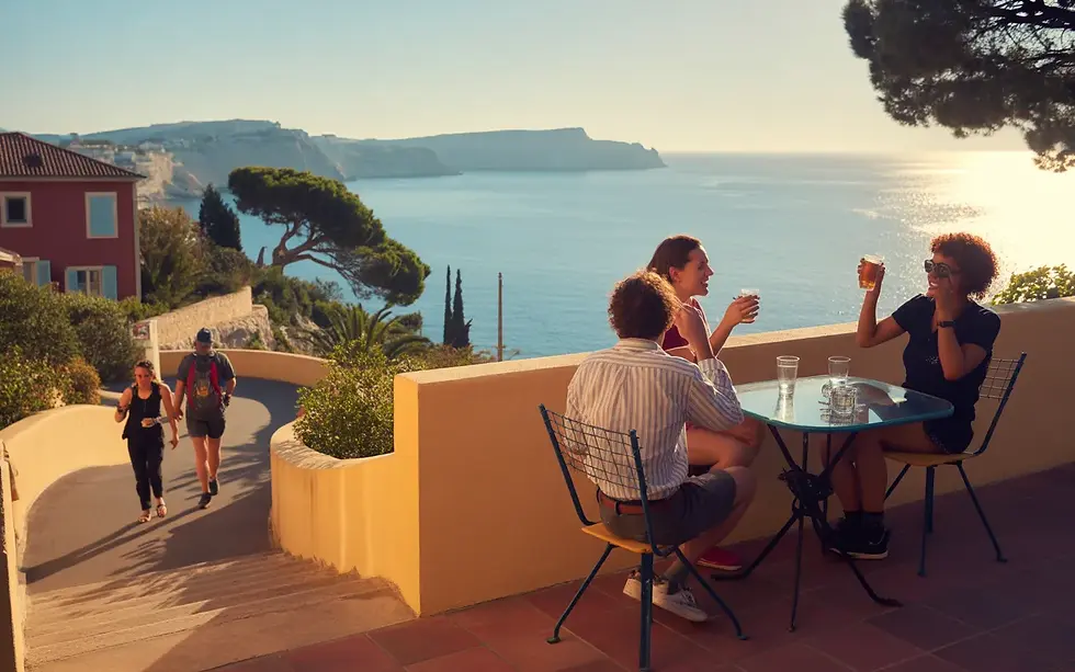 Vue panoramique en 16:9 d’une terrasse méditerranéenne conviviale dominant la côte, avec groupe de personnes discutant, randonneurs et joggeur sur un sentier descendant vers la mer, maisons aux toits rouges, pins parasols, criques rocheuses et calanques au loin, illustrant une activité week-end Marseille Est en plein air sous une douce lumière de fin d’après-midi.