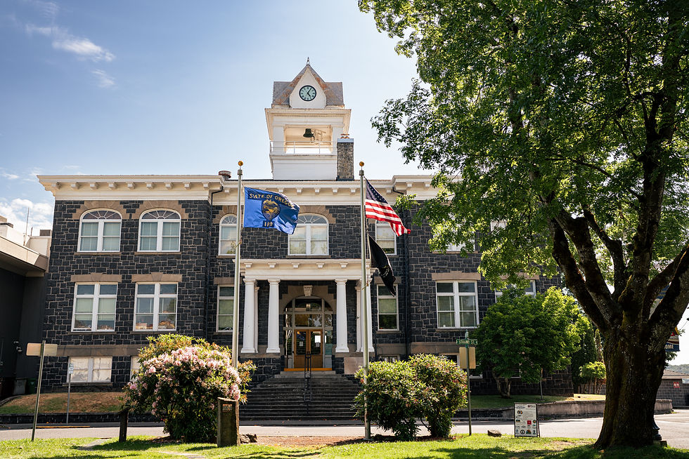 Historic St. Helens Columbia County courthouse with clock tower, Oregon flag, and U.S. flag in front. Surrounded by trees and bushes under a clear blue sky.