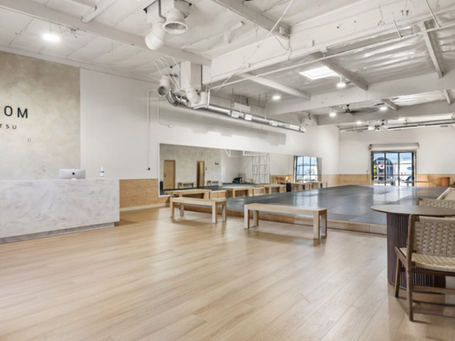 Interior view of Phenom Jiu Jitsu academy, featuring a modern and spacious environment with minimalist design. The space includes a marble reception desk, light wood flooring, natural lighting, and a mat area in the background set up for functional training.
