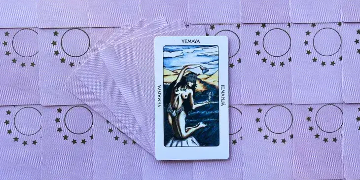 uma mulher branca semi nua segurando um espelho em uma mao e um pente em outra, usando uma coroa de conchas de joelho em uma pedra na beira de um rio ou mar.