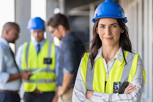 a project manager wears high-vis and a hard hat, behind her another project manager discusses the construction project with the clients
