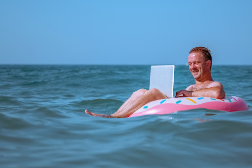 working from anywhere: a smiling man works at his laptop while floating in a rubber ring in the ocean