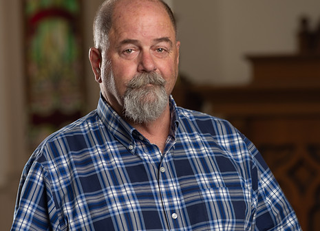 Man with beard in blue plaid shirt, blurred church background.