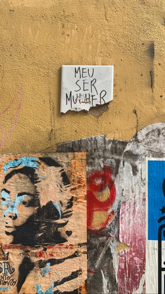 Azulejo branco com a frase MEU SER MULHER escrita com caneta preta, com a parte de baixo quebrada. O azulejo está aplicado em uma parede amarela, onde alguns outros desenhos estão colados na parte de baixo.