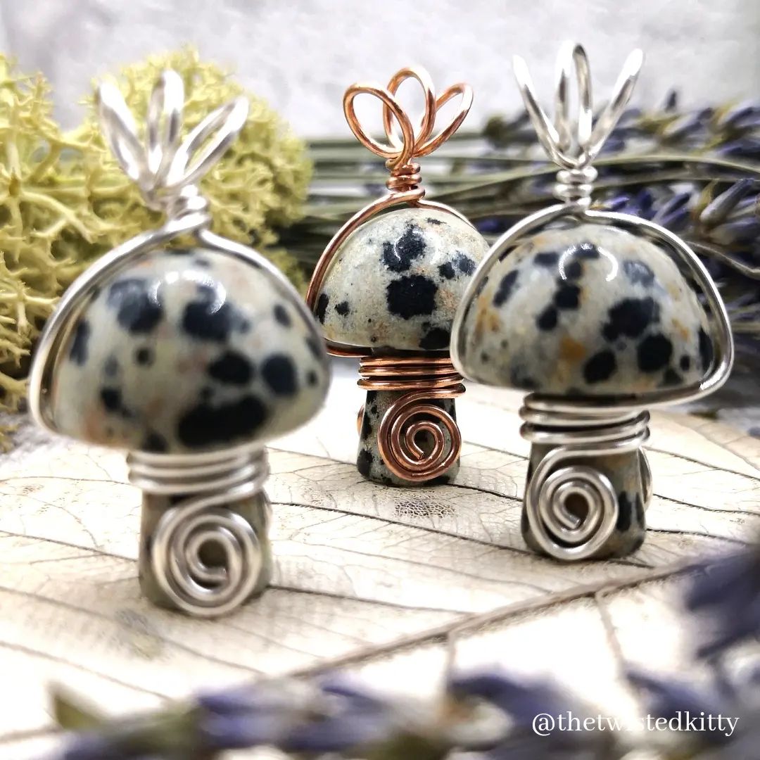 The image showcases three pendants shaped like mushrooms, crafted from speckled Dalmatian jasper stone and wire-wrapped in a spiral design. Each pendant features a cap and stem design, with swirling wire patterns around the stem and loops at the top for hanging. Two pendants are adorned with silver wire, while one is designed with copper wire. The background includes dried flowers and a textured surface, adding a rustic and artistic feel to the composition.