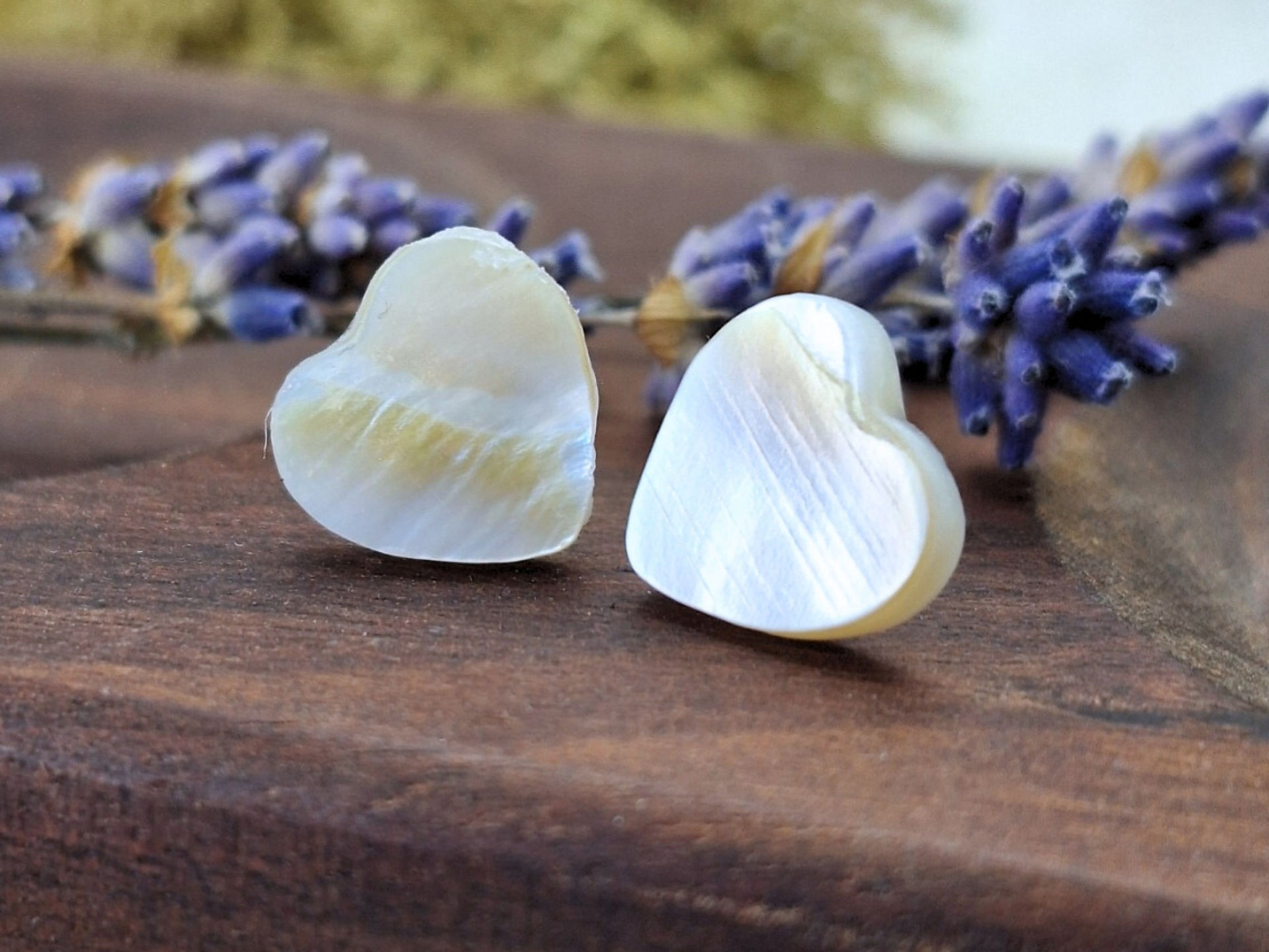 A pair of heart-shaped earrings crafted from mother-of-pearl sits on a textured wooden surface. The earrings display a smooth, iridescent finish with colours shifting from white to pale yellow. In the background, sprigs of lavender add a natural, rustic touch, complementing the artisanal and elegant feel of the earrings.