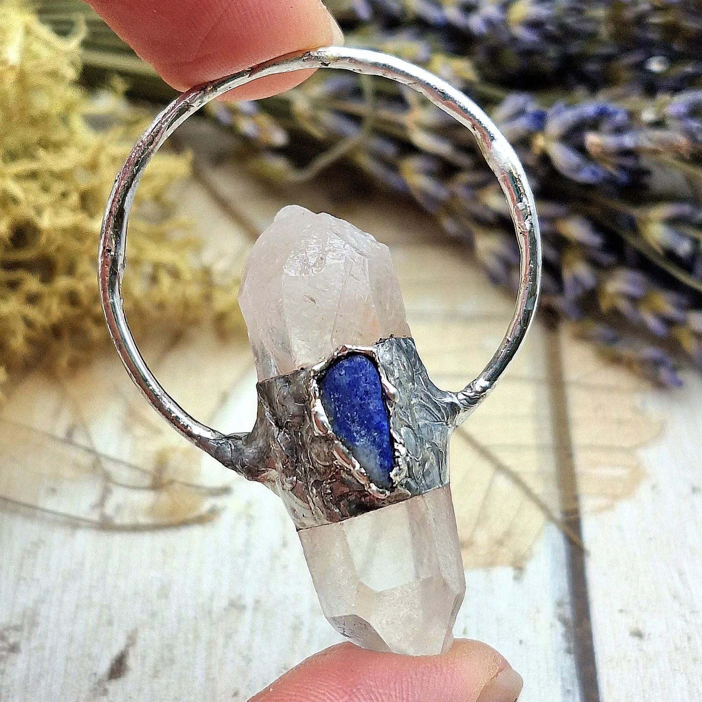 A hand gently holds a pendant made from a clear quartz crystal with a rough texture. The crystal is vertically set within a circular silver band, which features a small embedded lapis lazuli gemstone for added detail. Surrounding the pendant are dried flowers and sprigs of lavender, creating a natural and rustic backdrop that enhances the delicate and artisanal design of the jewellery.