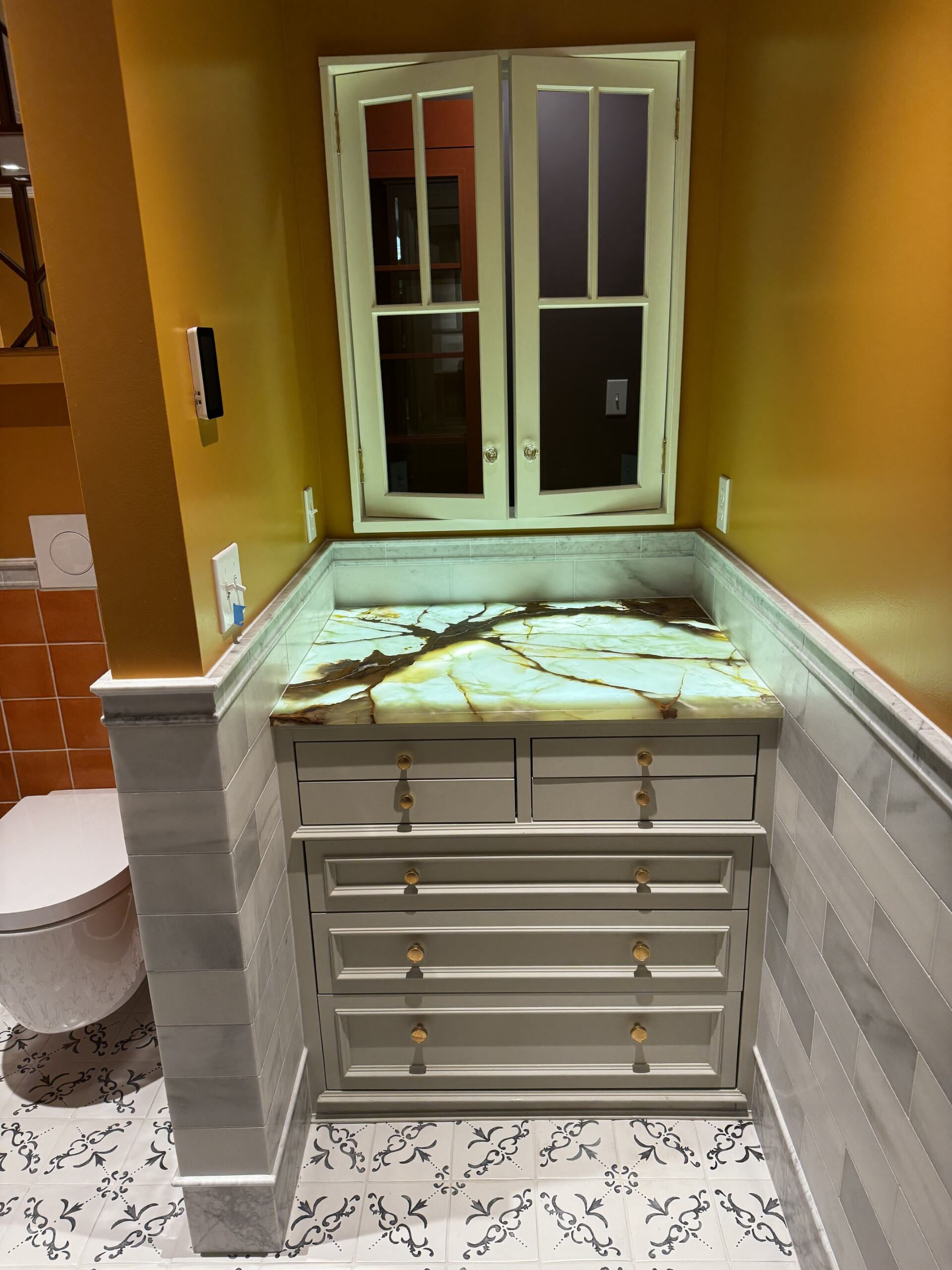 "Stylish bathroom corner featuring custom cabinetry with a stunning onyx countertop that glows subtly under lighting. The space includes elegant marble tile work, a modern wall-mounted toilet, and intricate patterned flooring.