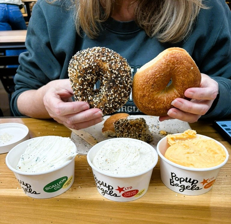 A woman holds an everything and plain bagel in front of 3 tubs of schmear from Popup Bagels