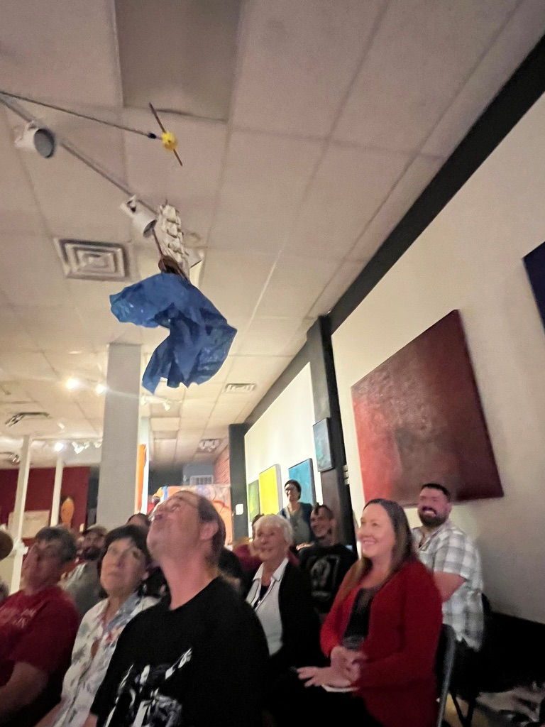An audience is entertained in an art gallery during a humorous art talk as a ship puppet floats over their heads.