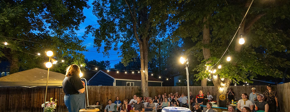 An audience laughs at the professional comedian on stage in an outdoor setting at dusk with whimsical lighting.