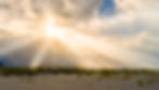 Sunbeams break through clouds over a sandy dune with beach grass, creating a warm, serene coastal scene.