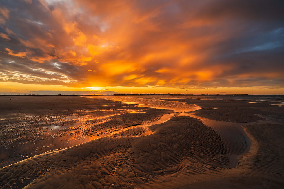 Glowing orange sunset over tidal flats, with rippled wet sand and dramatic clouds stretching across the horizon.