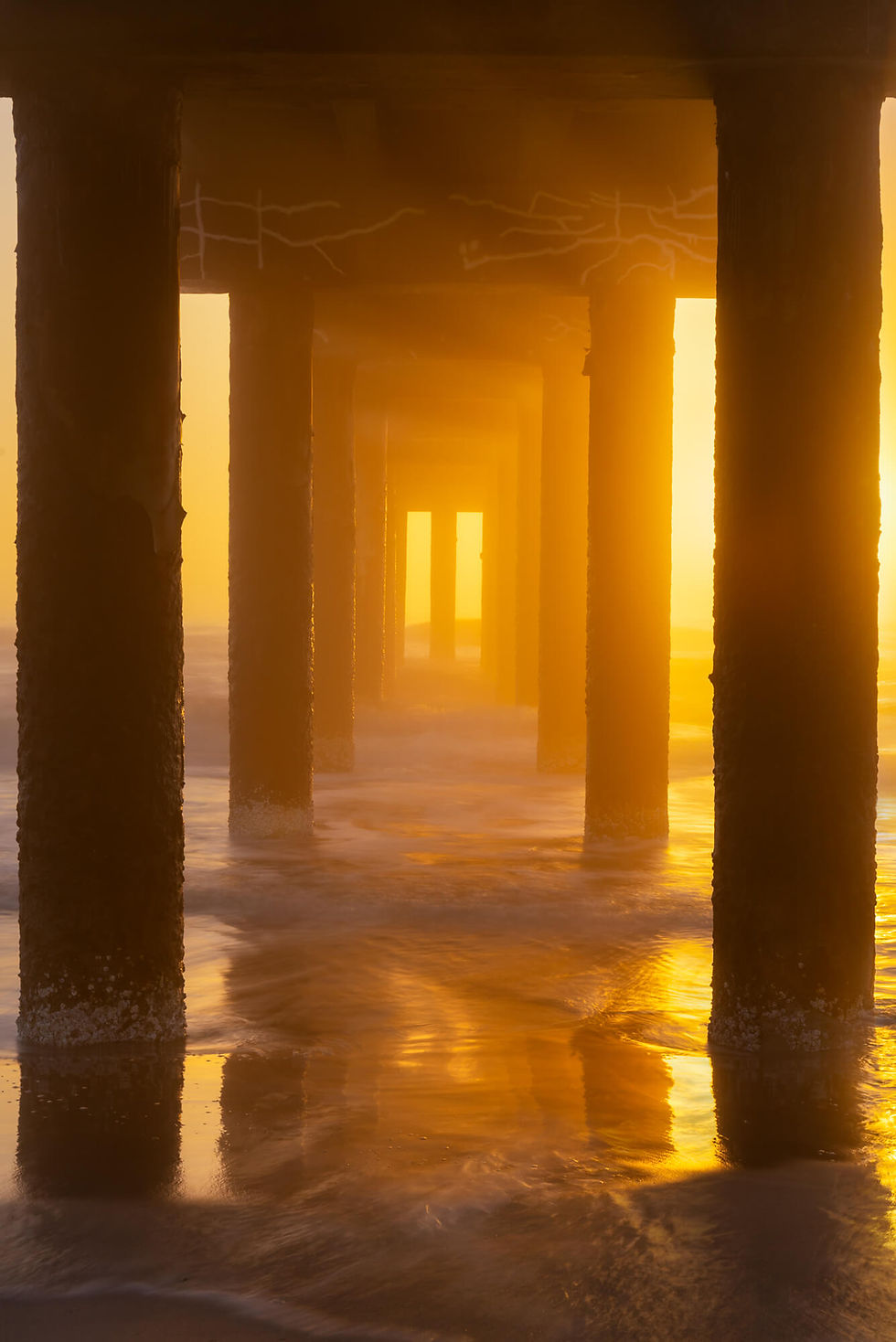Golden sunrise under the St Augustine pier, with dark pilings and waves washing through the misty ocean beneath. Light rays shine through thick fog.
