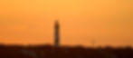 The St. Augustine lighthouse silhouetted against an orange sunset sky above dark treetops and houses, with The Great Cross nearby.