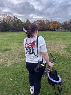 A person with long hair, wearing a white T-shirt with a graphic design, stands in a green park holding a camera and a tripod. Trees with autumn leaves are visible in the background.