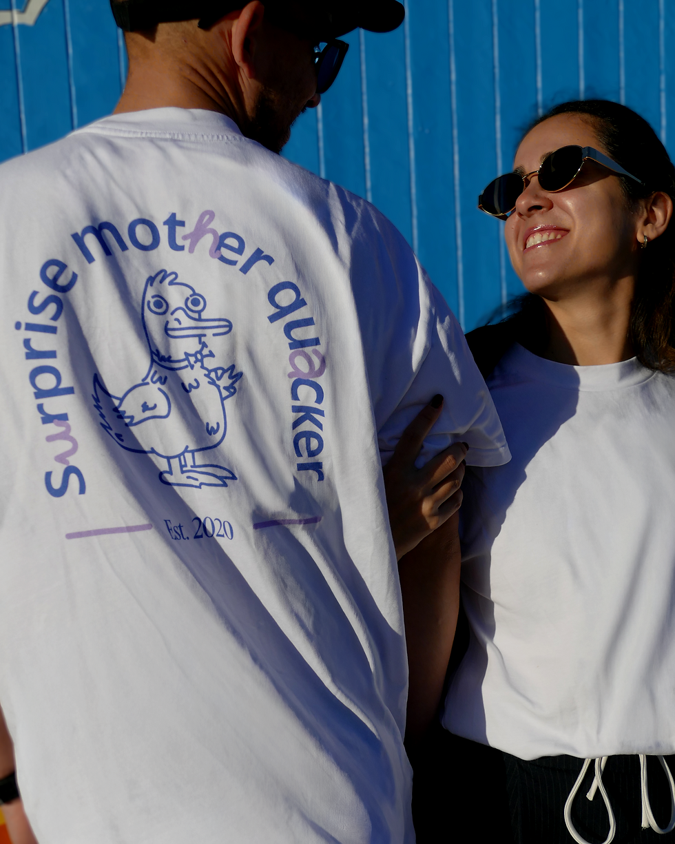 A person wearing a white t-shirt with a graphic of a duck and the text "Surprise mother quacker". Another person in a white shirt stands beside them, both smiling against a blue background.