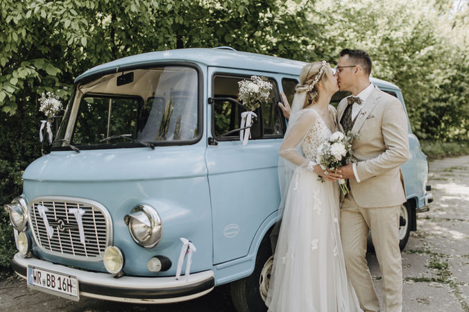 Brautpaar küsst sich vor blauem Oldtimer-Bus vor Bäumen. softgoldenpictures