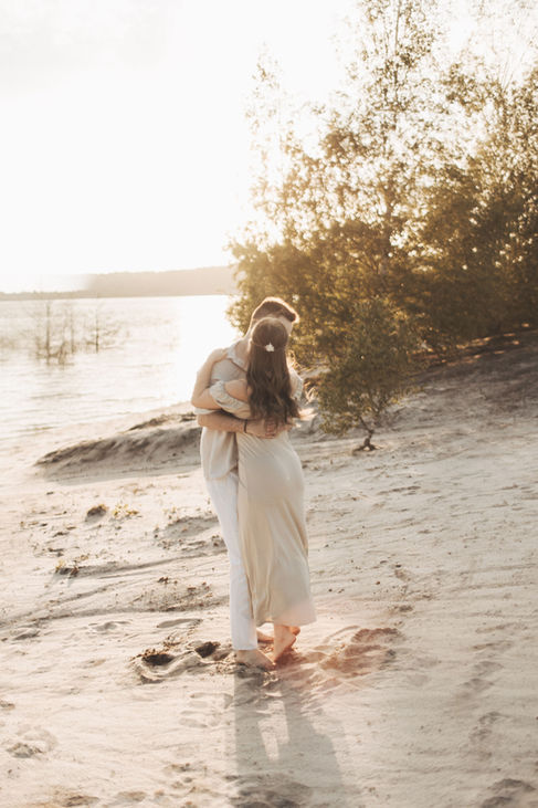 Paar umarmt sich am Strand, im Hintergrund Bäume und mildes Sonnenlicht.