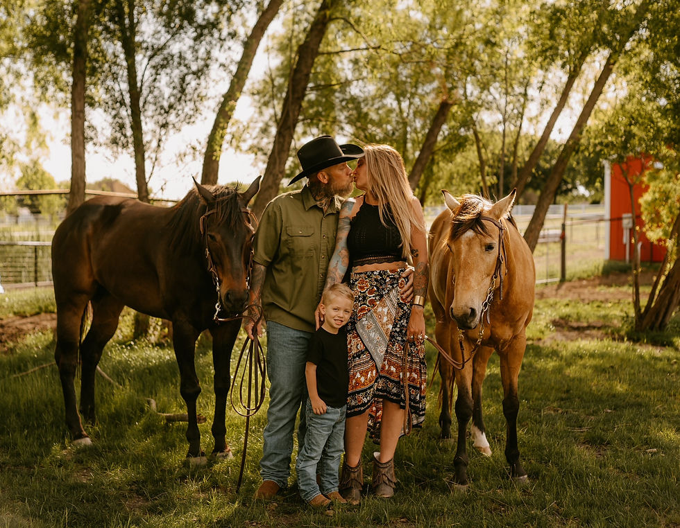 A couple shares a tender kiss with their child at their feet, surrounded by two gentle horses in a picturesque outdoor setting.