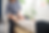 A therapist in a blue top works with a client lying on a treatment table, gently supporting the client's arm and head in a bright, plant-filled therapy room.