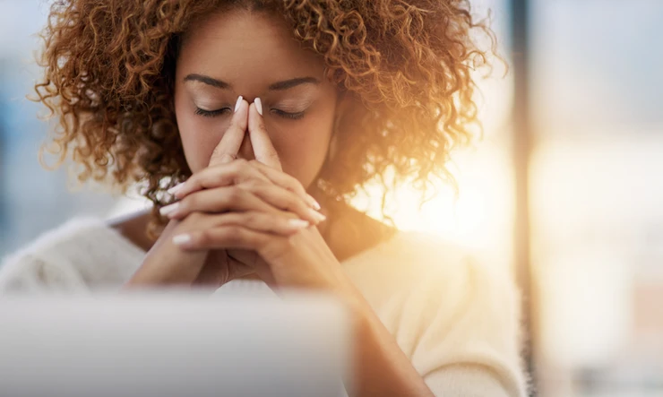A woman sits behind laptop with eyes closed and fingers steepled in front of her mouth