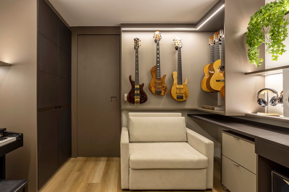 Espaço de escritório com uma decoração focada em instrumentos musicais. Na parede à direita, há uma coleção de guitarras e baixos pendurados em suportes, com uma iluminação embutida que destaca os instrumentos. Em frente à parede de instrumentos, há uma poltrona bege. À esquerda da poltrona, há uma mesa de trabalho com gavetas e prateleiras, incluindo fones de ouvido expostos em suportes. O ambiente é decorado com tons neutros e possui iluminação embutida, proporcionando uma atmosfera acolhedora e organizada.