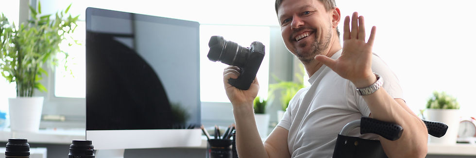 Man sitting in wheelchair smiling and waving to camera with 35mm in other hand.