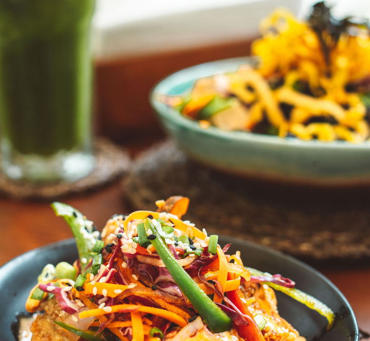 Crispy golden chicken topped with colorful shredded vegetables, peppers, green onions, and sesame seeds, served on a black plate.