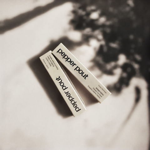 AI-generated product mockup of two boxes of lip gloss, shown on a beige surface with soft tree-shadow lighting