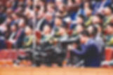 A cameraman films an event in a crowded auditorium. Attendees in military and formal attire sit attentively. The setting is formal.