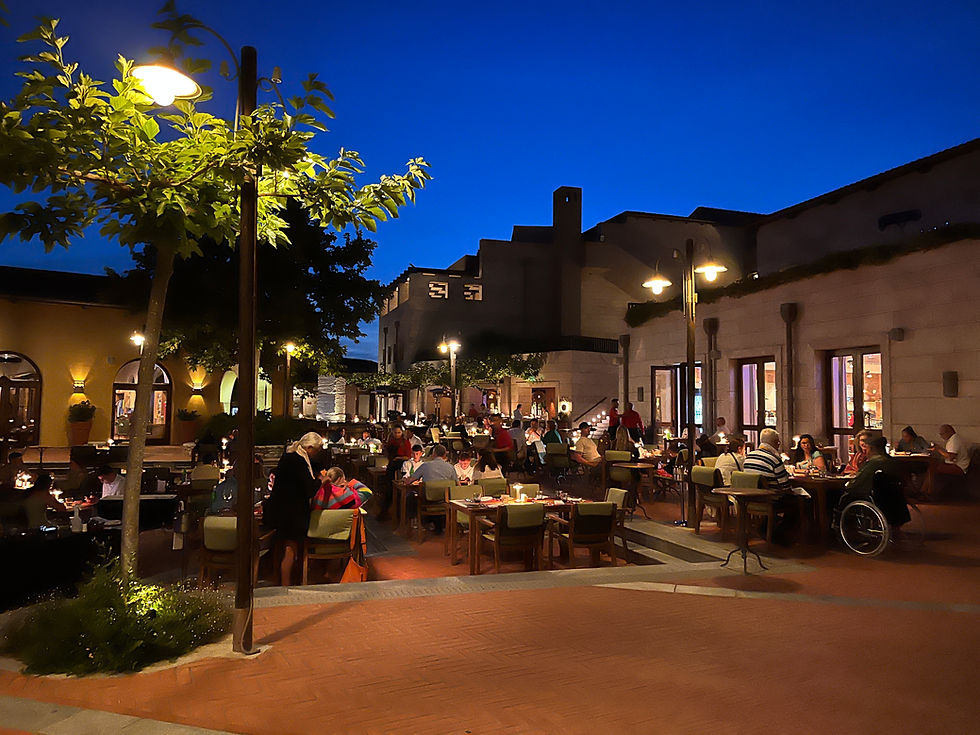 A dark image of an outdoor dining area. There are buildings on the right hand side, leading out to an open pavilion that is set out with many dining tables at which people are sat. The area is split on two levels, with a few steps leading to the lower level. There is one small tree, and some streetlamps lighting the scene.