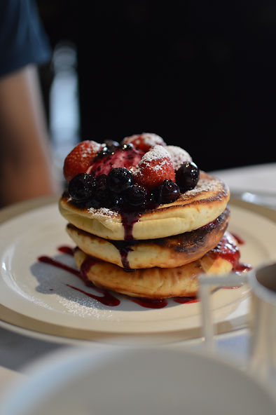 An image of a stack of three big pancakes on a white plate with a light brown rim. The pancakes have blueberries and strawberries on top with a dusting of icing sugar. The pancakes have been drizzled with a dark purple fruit sauce.