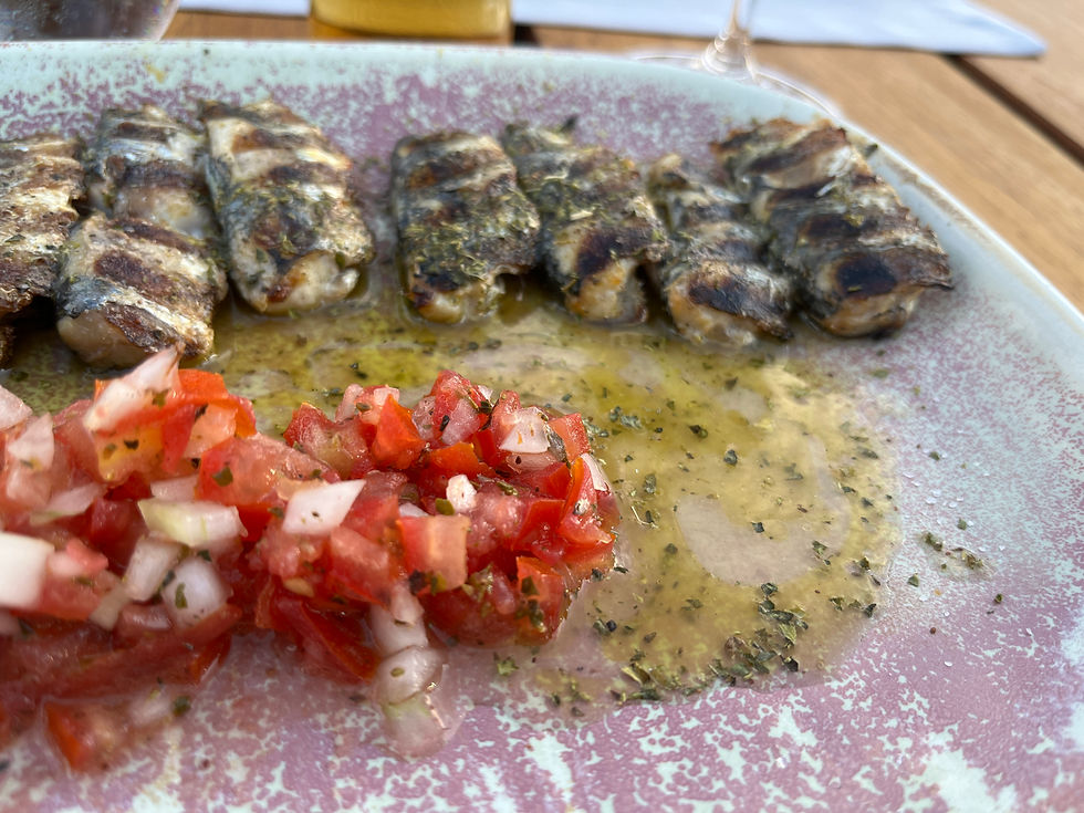 A reddish-blue plate on a wooden table, containing a line of small rectangular pieces of charred mackerel sitting in some herby olive oil. There is a mixture of chopped tomatoes and onions in front of the fish.