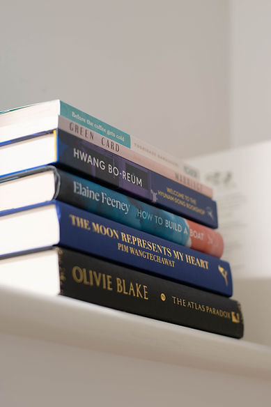 An image of a stack of books at an angle. The book titles displayed are: 'Before the coffee gets cold', 'Green Card Marriage', Welcome to the Hyunam-Dong Bookshop', How to Build a Boat', 'The Moon Represents My Heart' and 'The Atlas Paradox'.
The colours of the books include black, navy blue, deep purple, turquoise and pink, pale pink, and light blue.