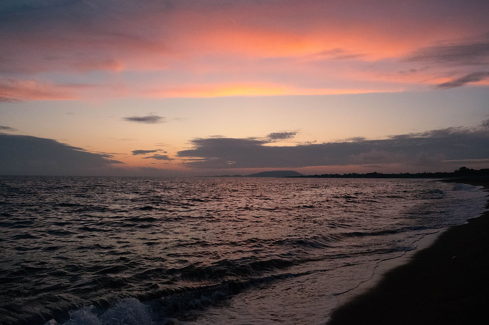 A photo of a beach at sunset. The sea is dark and wavy, taking up the lower half of the image. THe beach is slightly in view on the right but is totally dark. The upper half of the image is the sky, there are dark grey clouds closer to the sea and some pale blue sky in the middle. Deep pink and orange clouds fill the upper part of the sky.