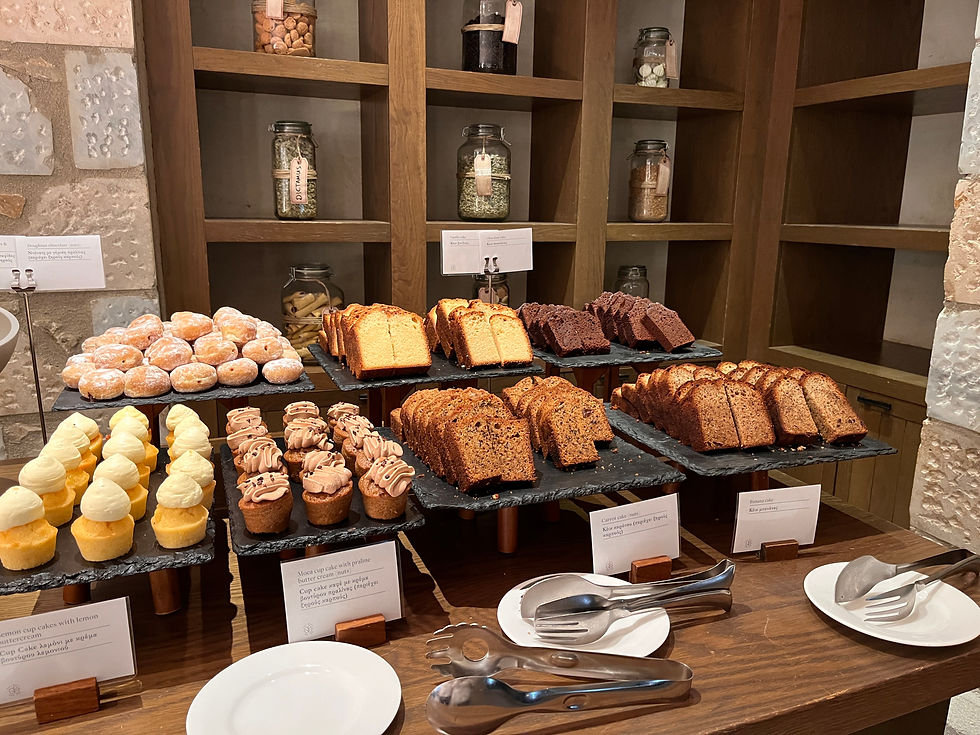 In the foreground of this image, there are slate platters of cakes and breads on a wooden table. Each platter has a white description card in front (text not clear). There are some white plates and serving tongs on the table. In the background, there are wooden shelves set into a stone wall. Each square-shaped shelf has a jar of something on it.
