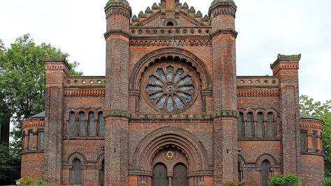 Historic brick building with ornate arched windows and a large circular rose window, surrounded by trees and greenery. Overcast sky.