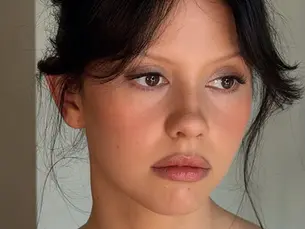 Young woman with dark hair looks to the side, in soft lighting against a plain background. A contemplative mood is conveyed.